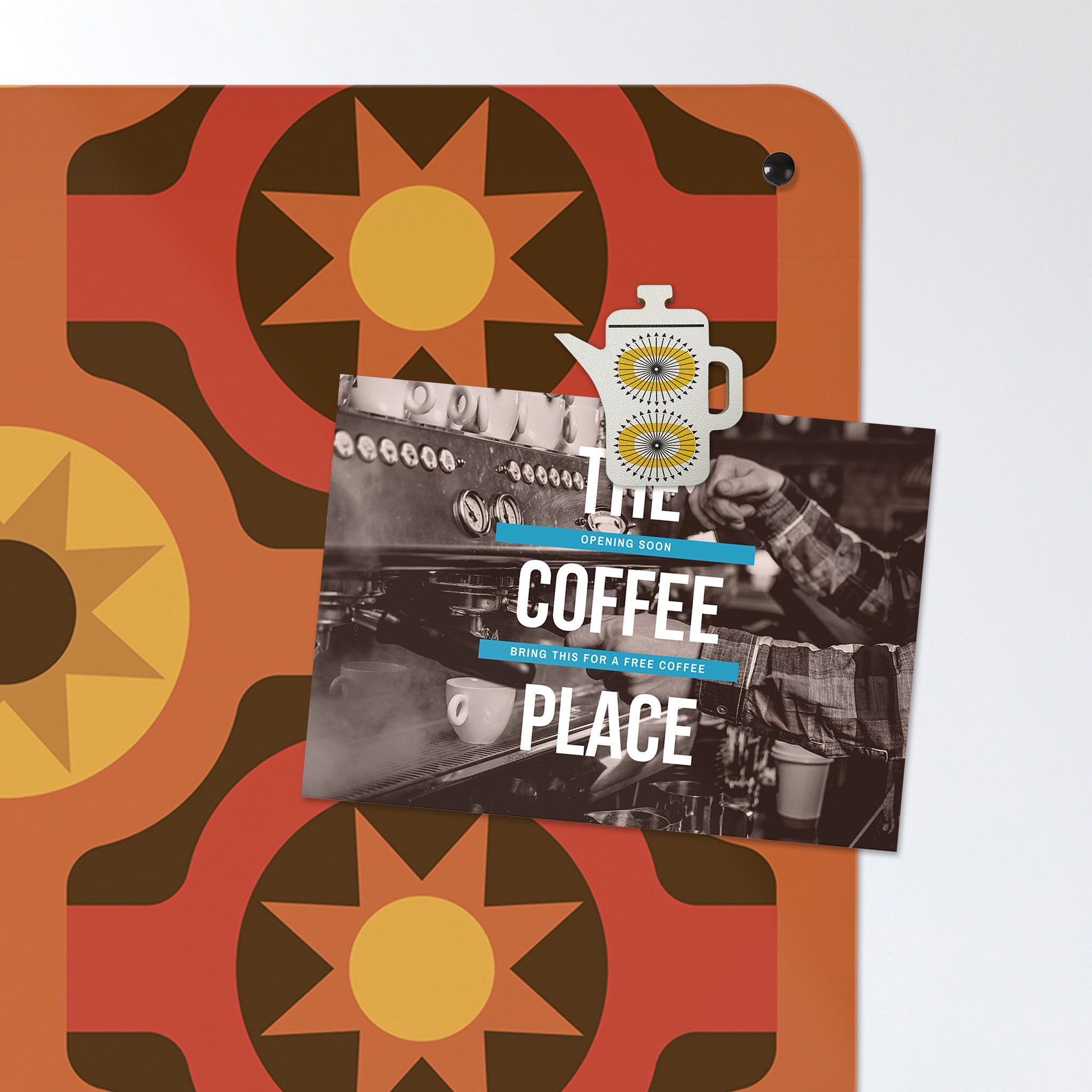 A corner metal of a large magnetic notice board showing a design of a  coffee pot with a quote that reads 'but first coffee' with a seventies style orange and brown pattern behind with a postcard attached with a coffee pot shaped magnet