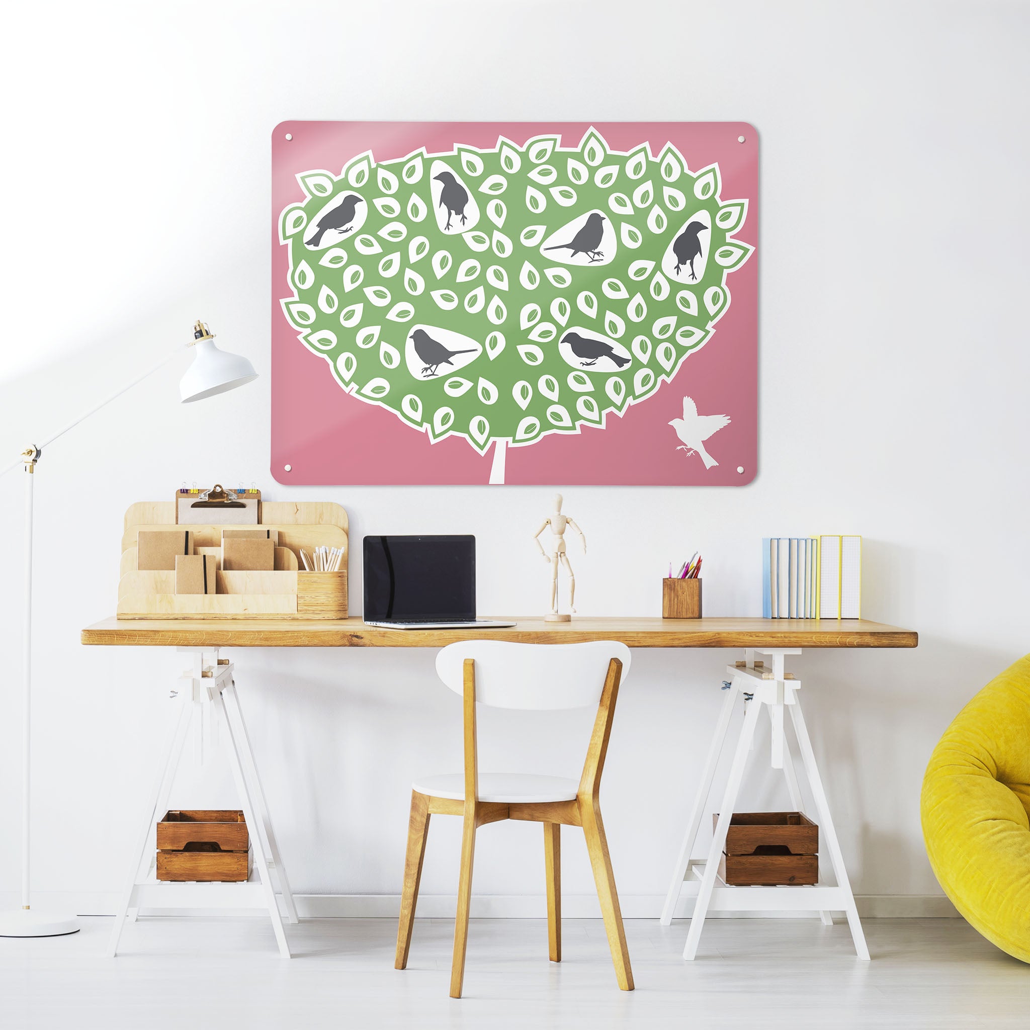 A desk in a workspace setting in a white interior with a magnetic metal wall art panel showing a black birds in a tree design