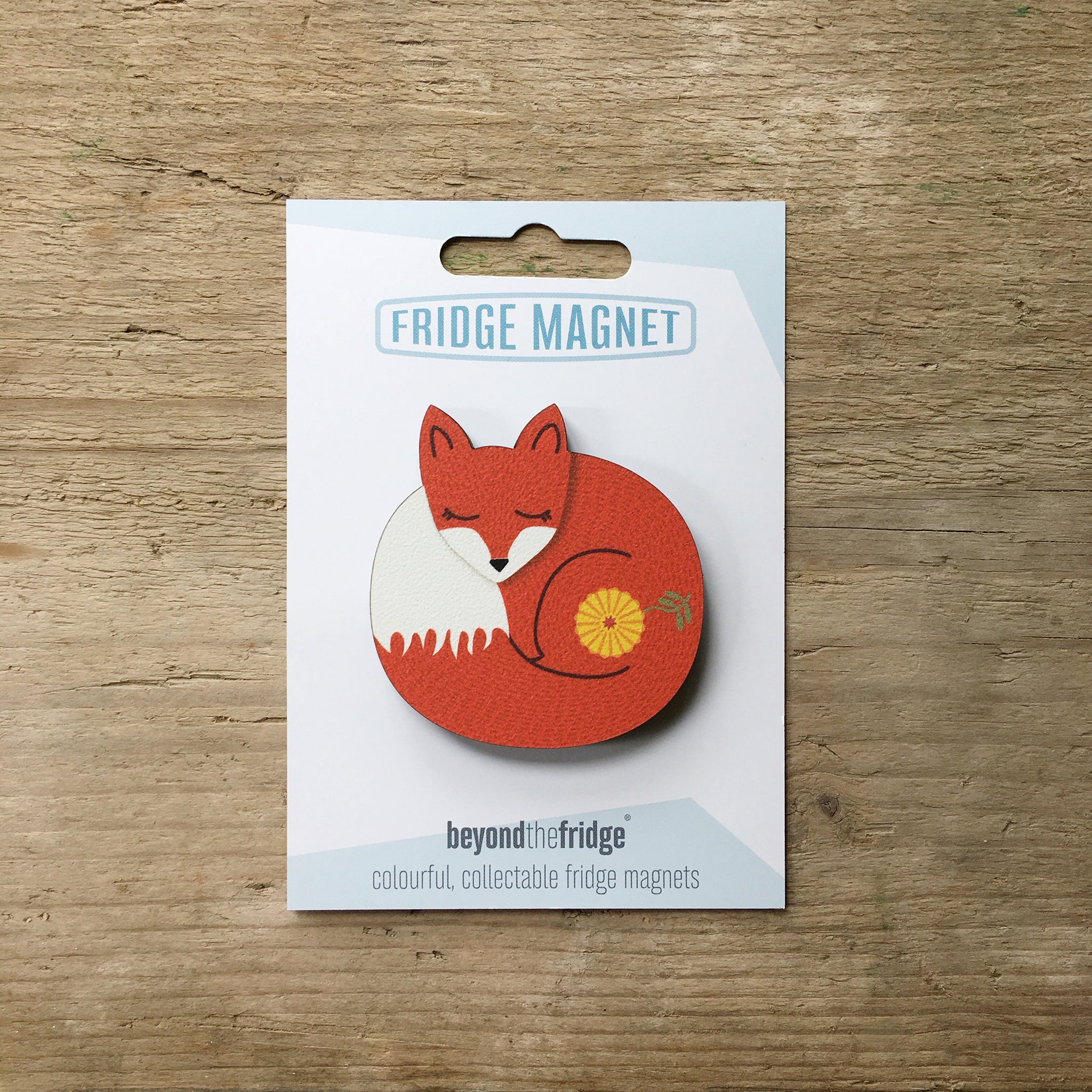 A sleeping fox design plywood fridge magnet by Beyond the Fridge in it’s pack on a wooden background