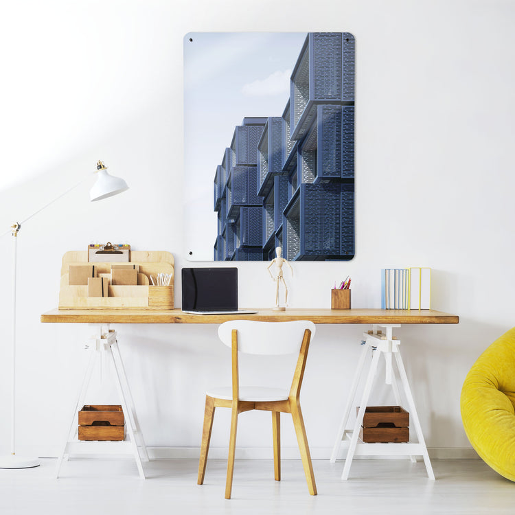 A desk in a workspace setting in a white interior with a magnetic metal wall art panel showing a photograph of a modern blue building