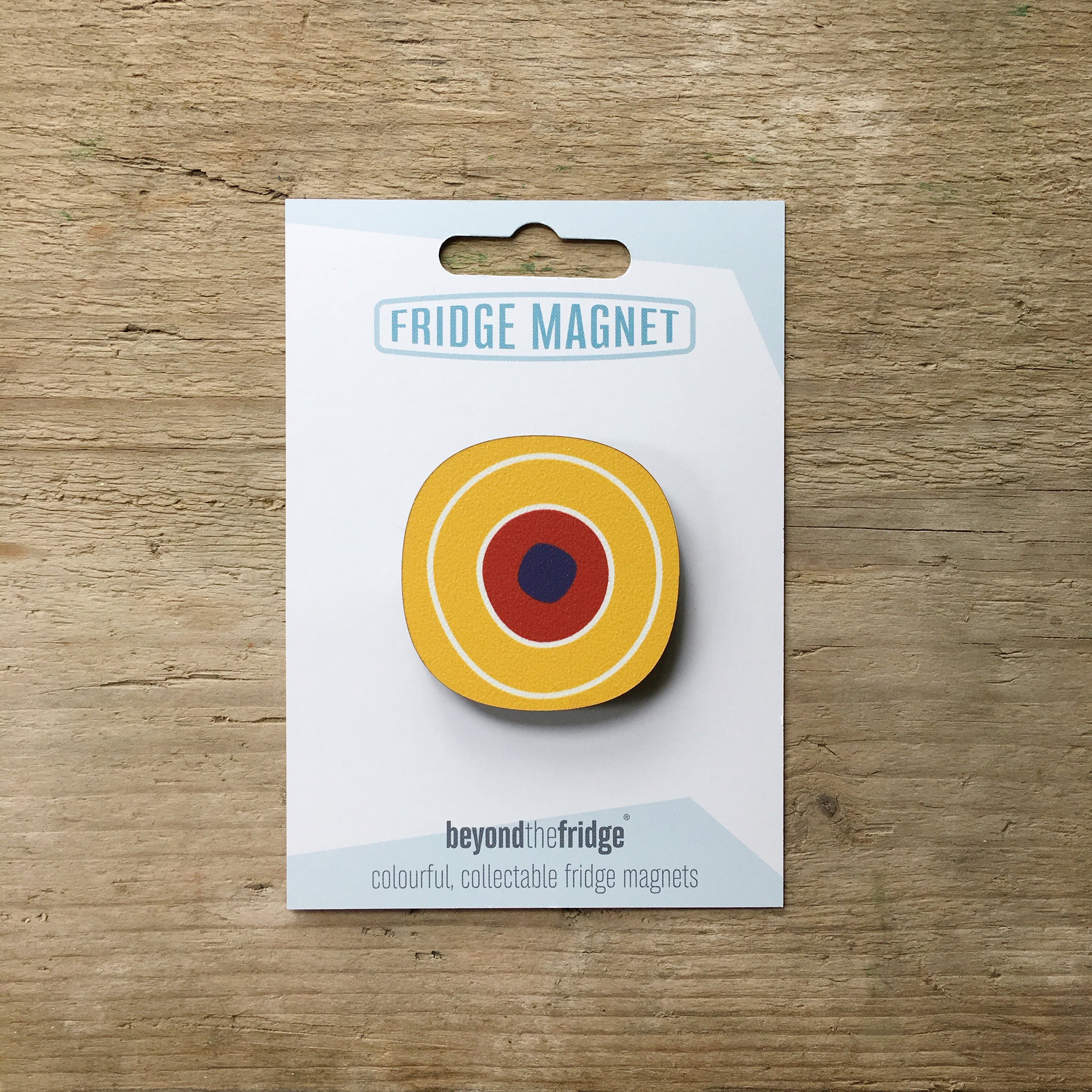 A yellow circle millefiori design plywood fridge magnet by Beyond the Fridge in it’s pack on a wooden background