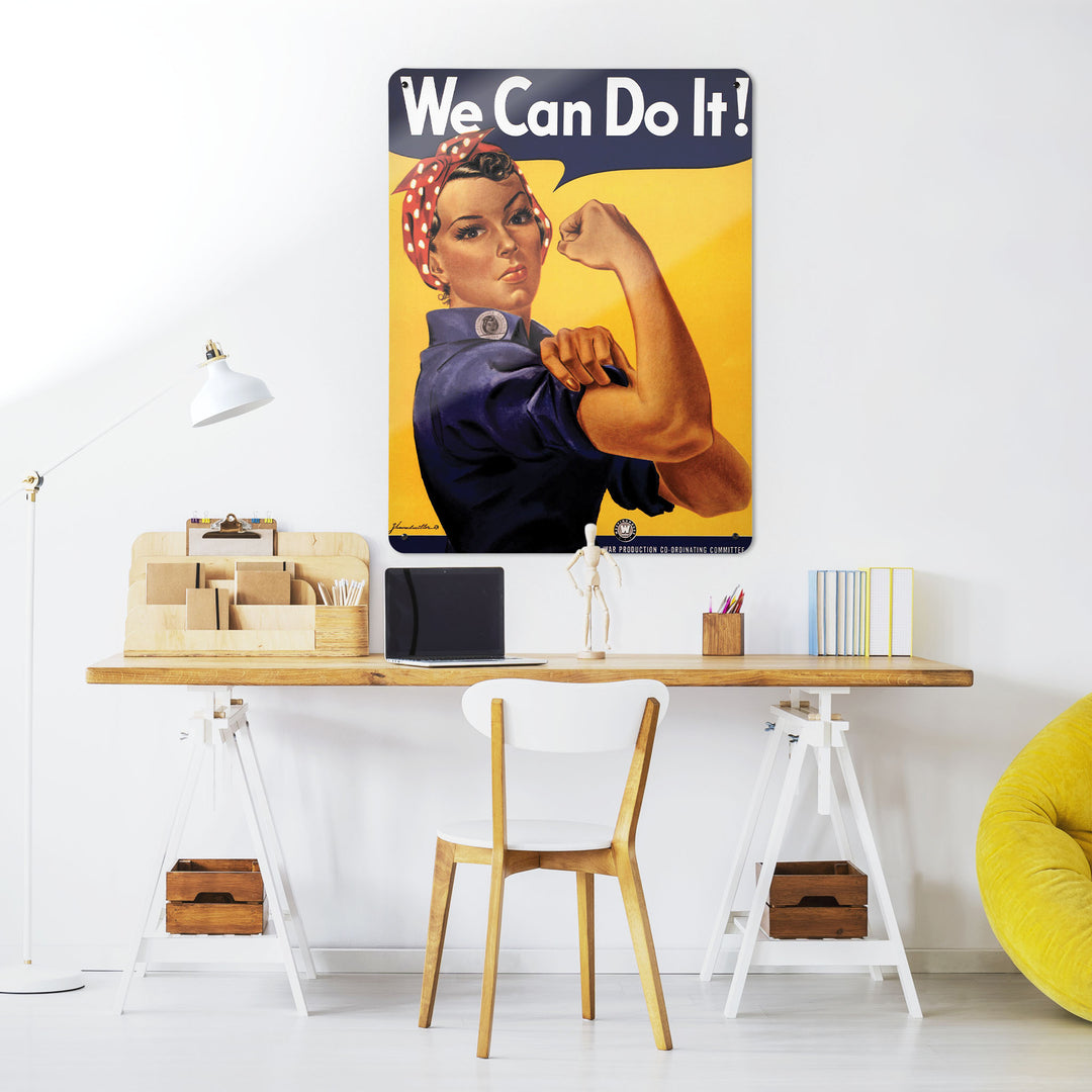 A desk in a workspace setting in a white interior with a magnetic metal wall art panel showing a wartime American retro poster of Rosie the riveter