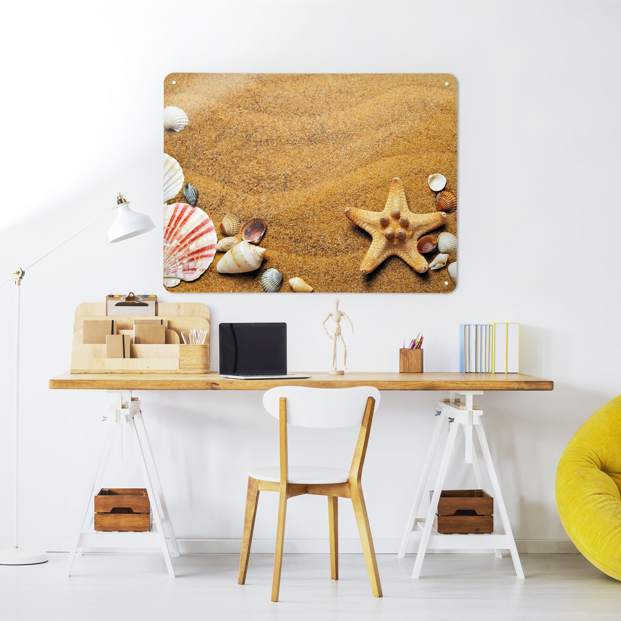 A desk in a workspace setting in a white interior with a magnetic metal wall art panel showing a sandy shore with seashells