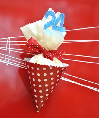 A hand crafted red polka dot paper cone with a gift inside and a blue glitter number 24 attached to a red magnetic board with a magnet