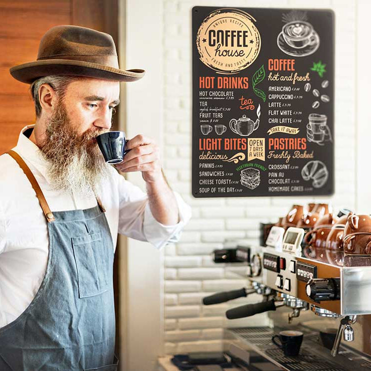 A barista with beard in apron drinking coffee in an artisan coffee shop with a magnetic board menu on the wall
