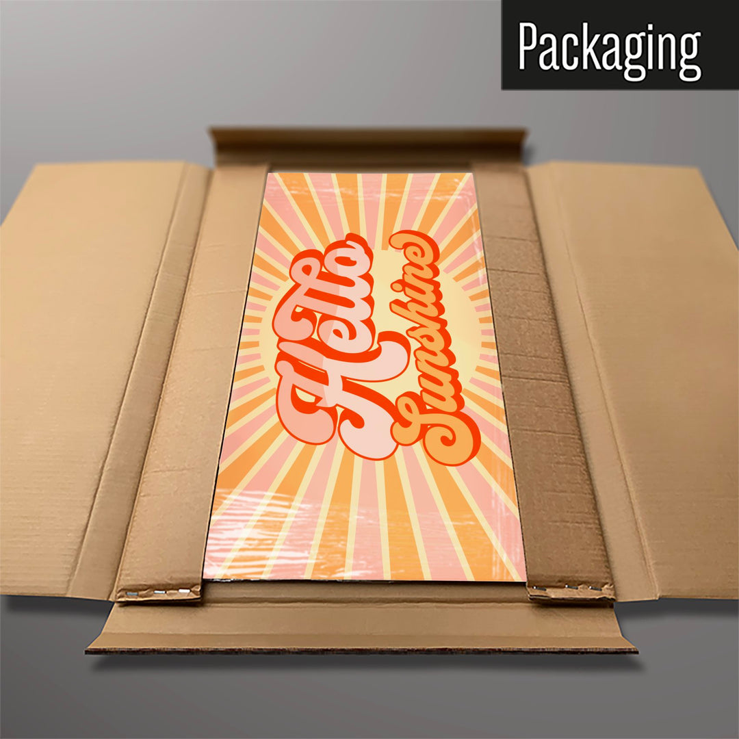 A large steel magnetic board with a print of a typographic quote design that reads Hello Sunshine with a sunburst in pink and orange colours in cardboard packaging