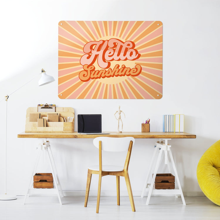 A large steel magnetic board with a print of a typographic quote design that reads Hello Sunshine with a sunburst in pink and orange colours in a workspace setting with a desk and chair