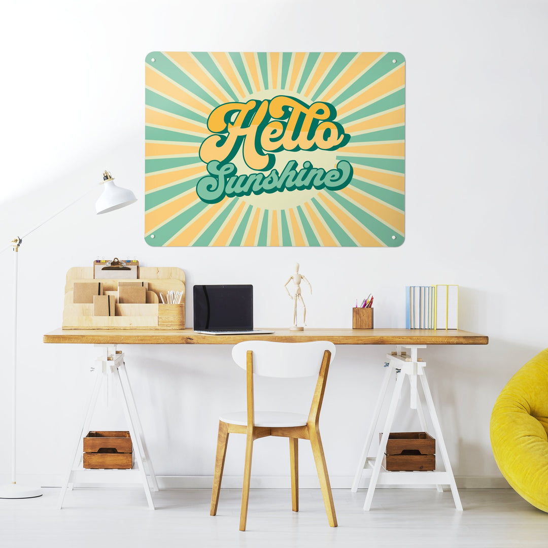 A large steel magnetic board with a print of a typographic quote design that reads Hello Sunshine with a sunburst in aqua and yellow colours in a workspace setting with a desk and chair