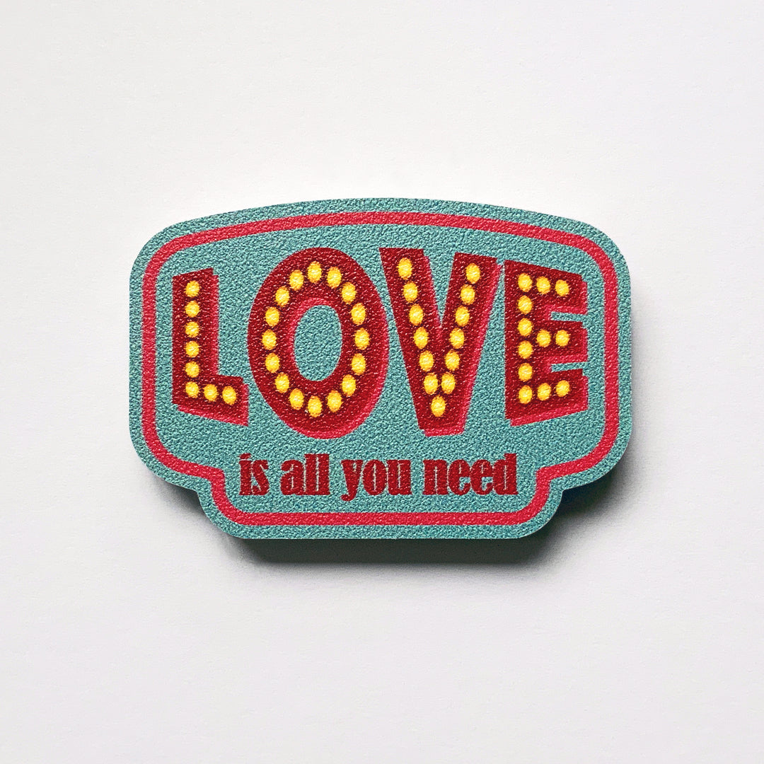 A vintage label shaped plywood fridge magnet by Beyond the Fridge with type that says love is all you need