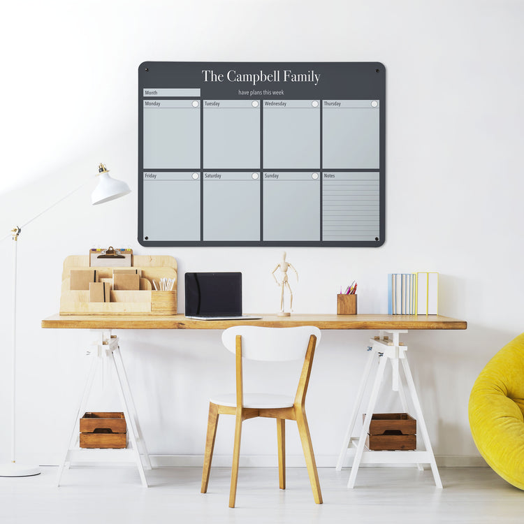 A desk in a workspace setting in a white interior with a dry wipe magnetic metal wall art panel showing a weekly planner design to personalise in grey landscape format