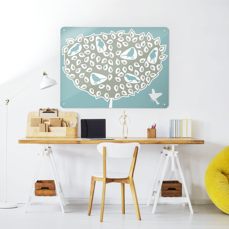 A desk in a workspace setting in a white interior with a magnetic metal wall art panel showing a blue birds in a tree design