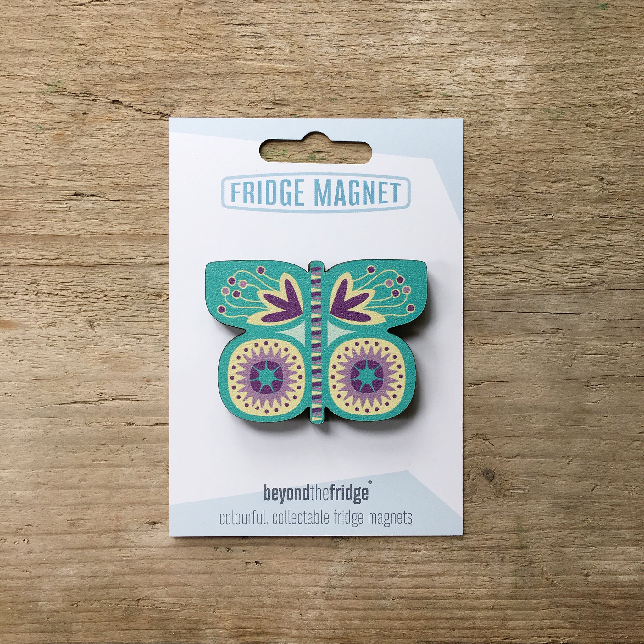 An aqua butterfly shaped plywood fridge magnet by Beyond the Fridge in it’s pack on a wooden background