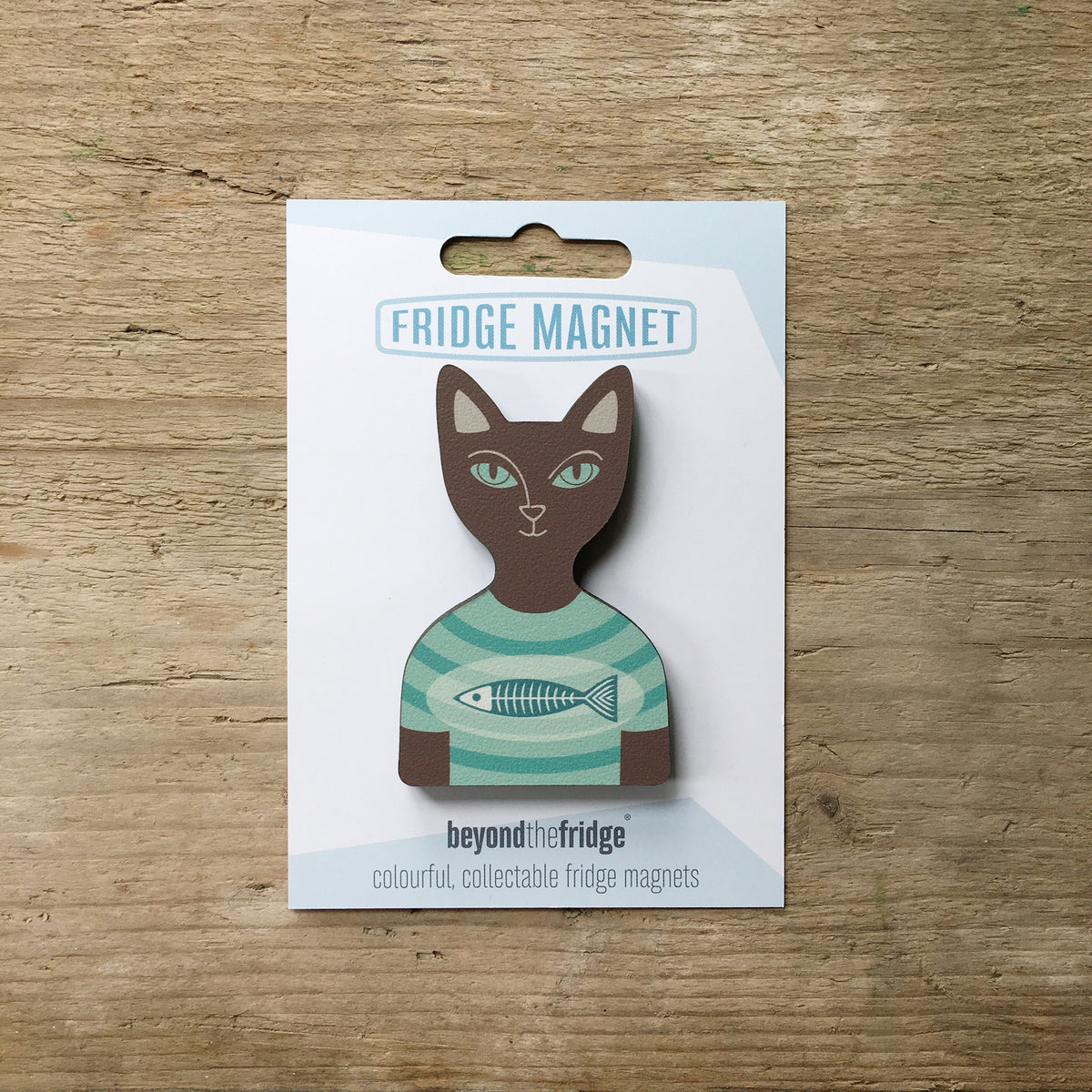 A brown cat in a t-shirt shaped plywood fridge magnet by Beyond the Fridge in it’s pack on a wooden background