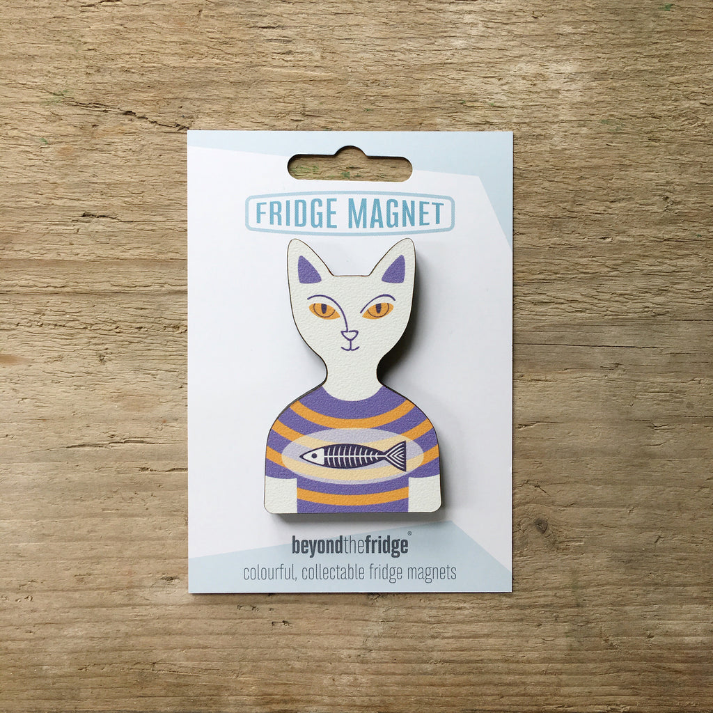A white cat in a t-shirt shaped plywood fridge magnet by Beyond the Fridge in it’s pack on a wooden background