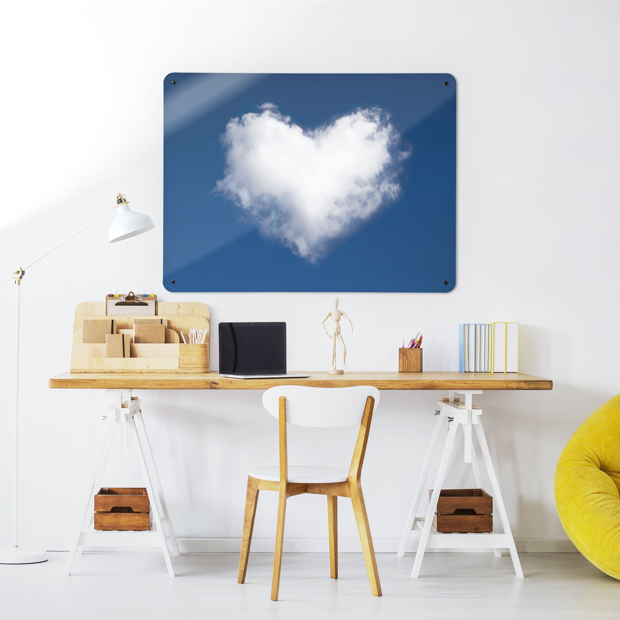 A desk in a workspace setting in a white interior with a magnetic metal wall art panel showing an image of a white cloud heart in a blue sky
