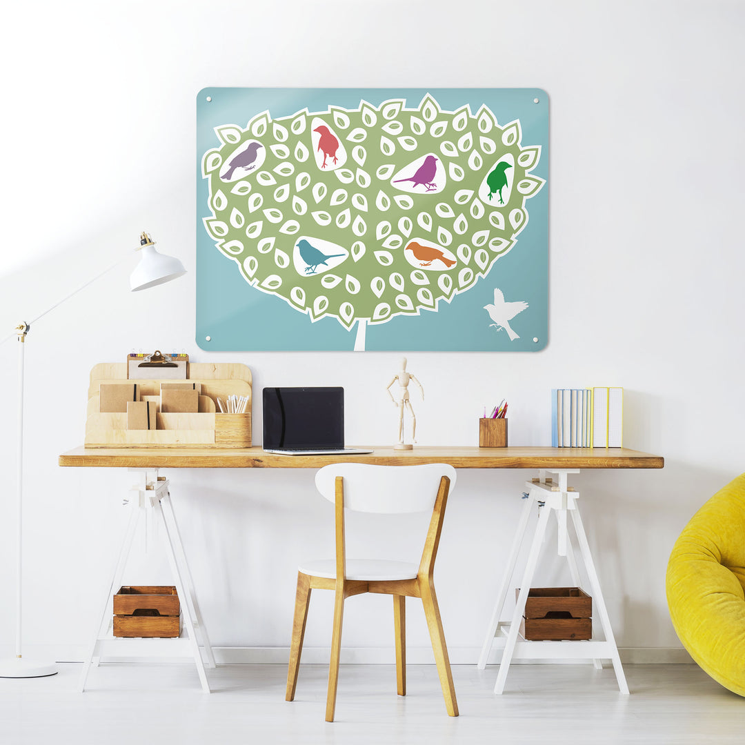 A desk in a workspace setting in a white interior with a magnetic metal wall art panel showing a coloured birds in a tree design