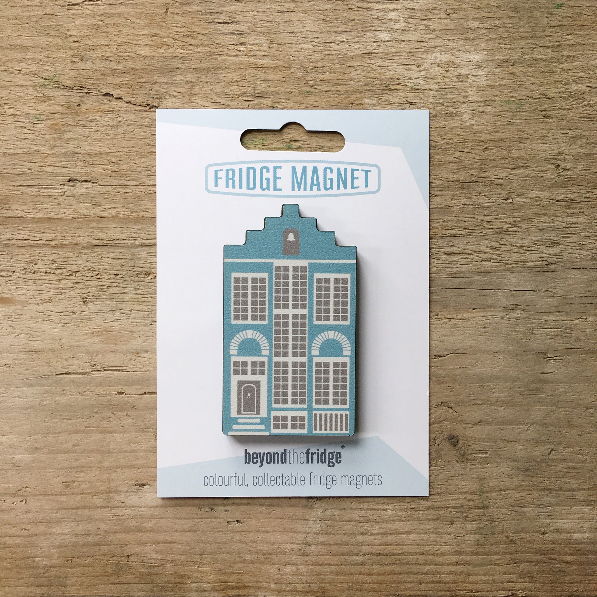 A blue Delft house shaped plywood fridge magnet by Beyond the Fridge in it’s pack on a wooden background