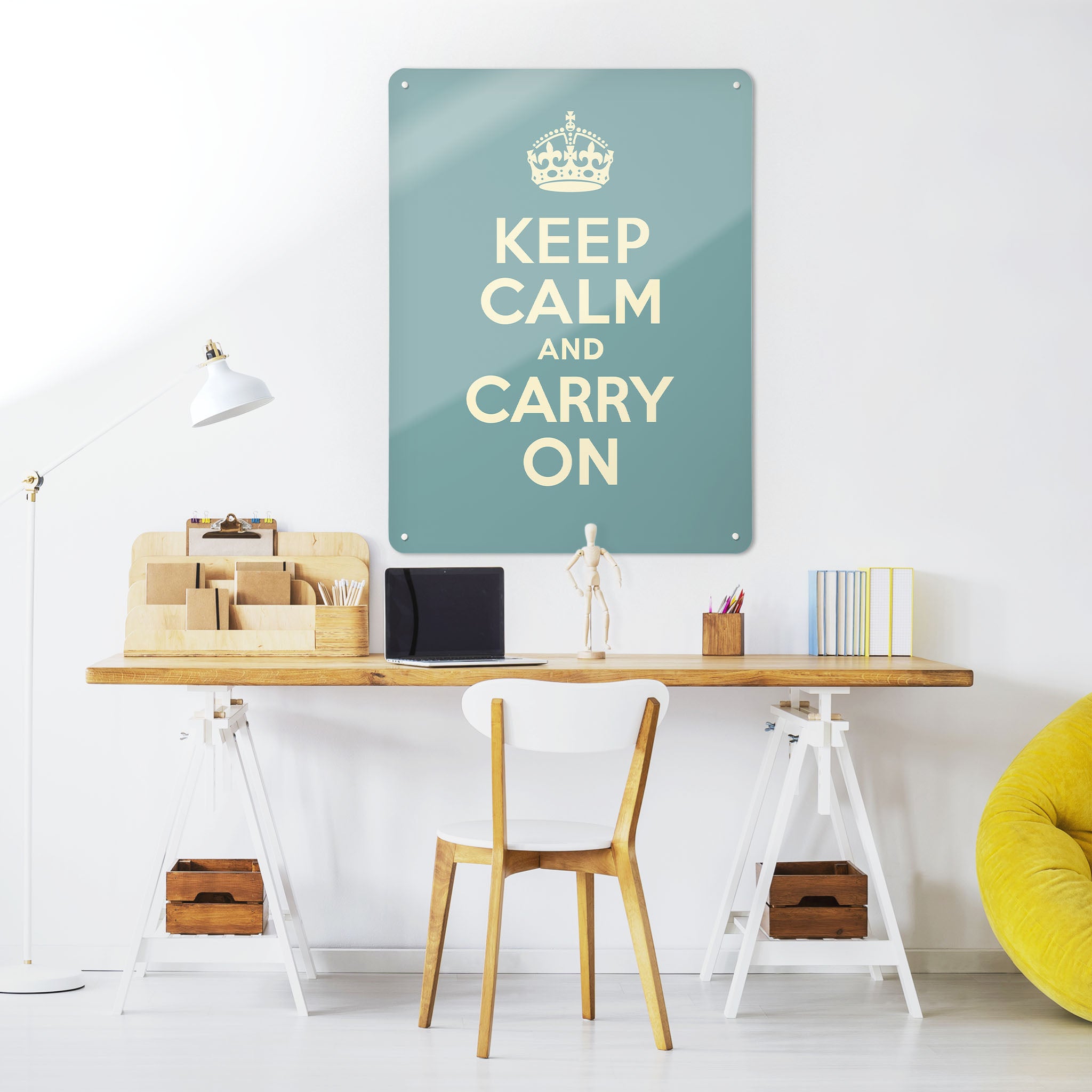 A desk in a workspace setting in a white interior with a magnetic metal wall art panel showing a vintage keep calm and carry on poster in blue
