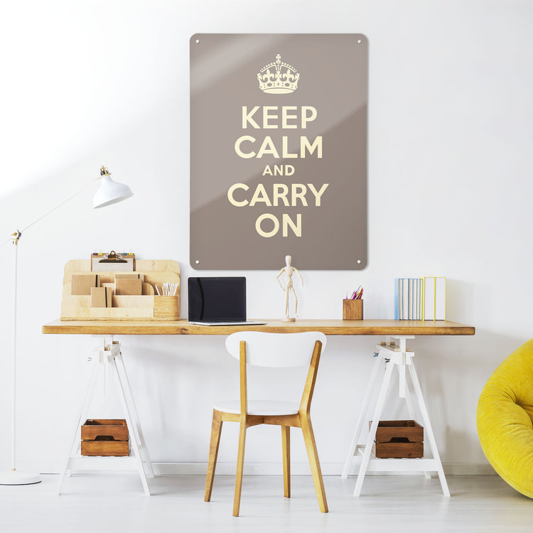 A desk in a workspace setting in a white interior with a magnetic metal wall art panel showing a vintage keep calm and carry on poster in brown