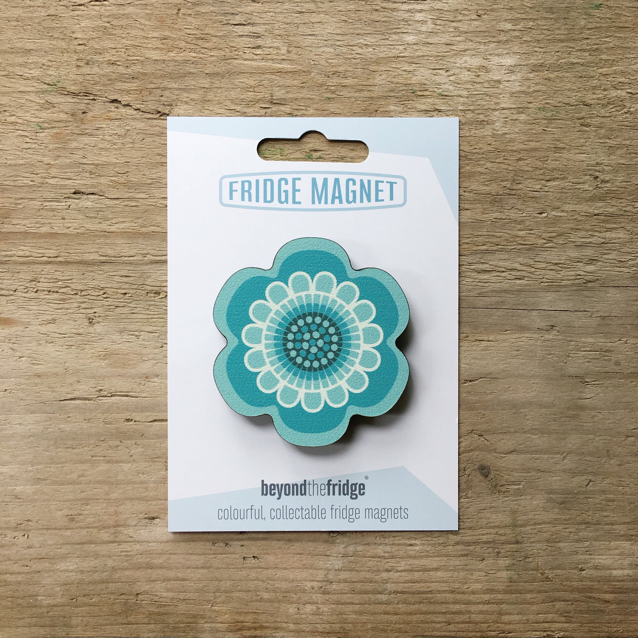A aqua flower design plywood fridge magnet by Beyond the Fridge in it’s pack on a wooden background