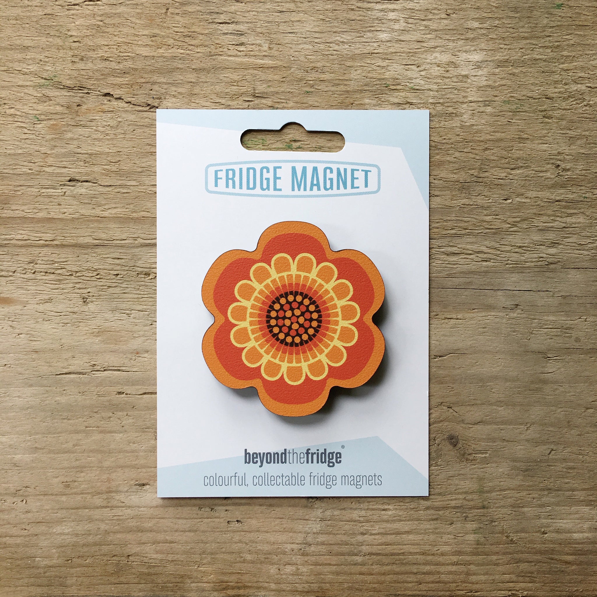 A orange flower design plywood fridge magnet by Beyond the Fridge in it’s pack on a wooden background