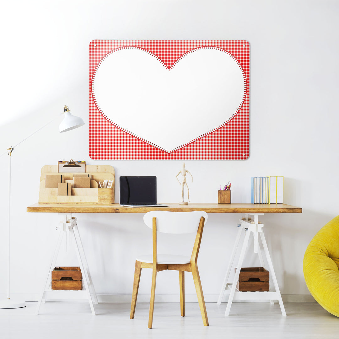A desk in a workspace setting in a white interior with a dry erase magnetic metal wall art panel showing a gingham heart design in red and white