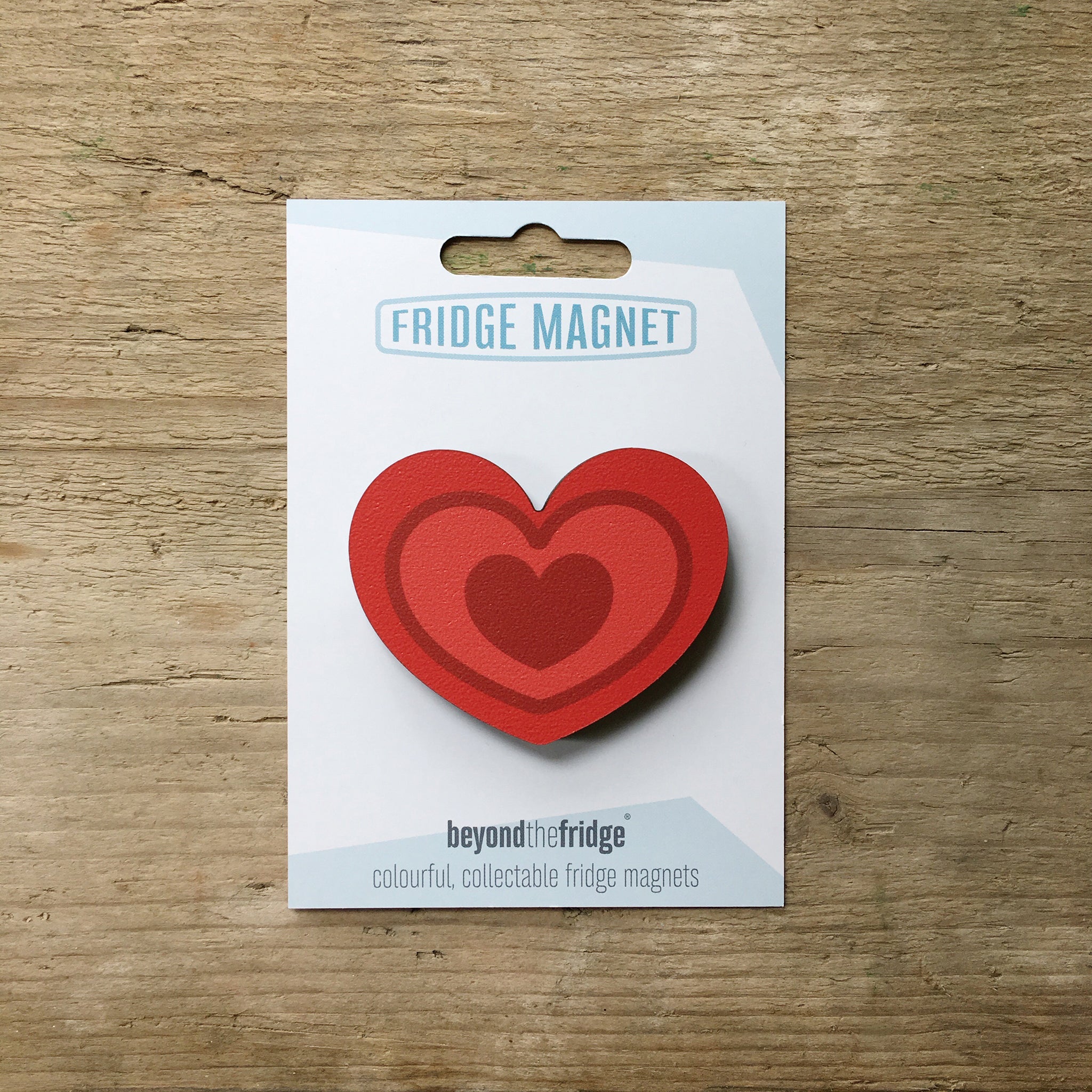 A red heart design plywood fridge magnet by Beyond the Fridge in it’s pack on a wooden background