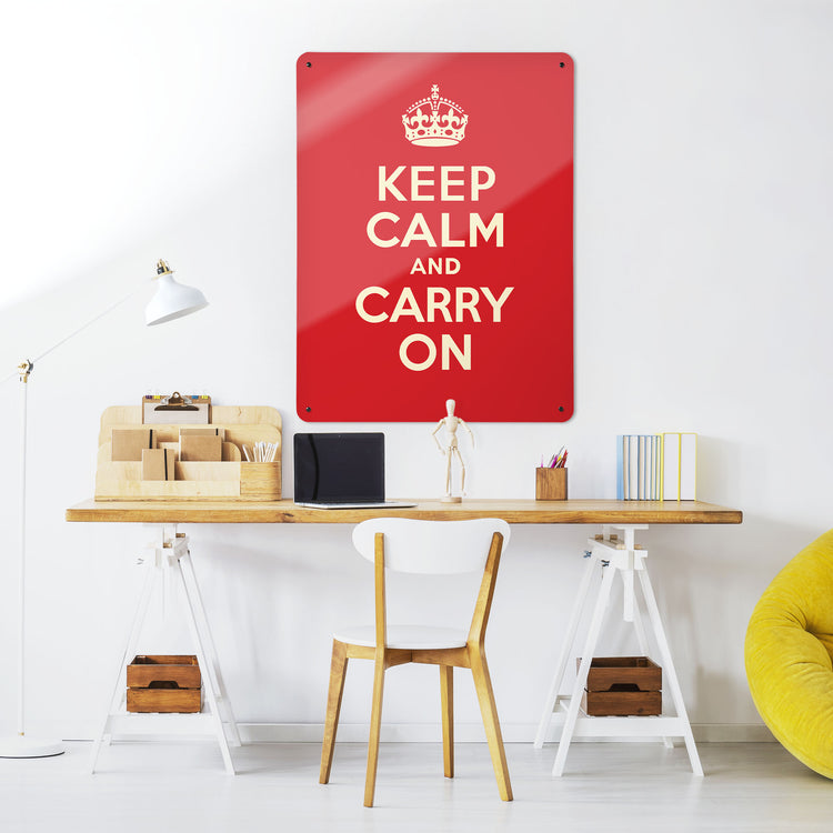 A desk in a workspace setting in a white interior with a magnetic metal wall art panel showing a red keep calm and carry on vintage poster