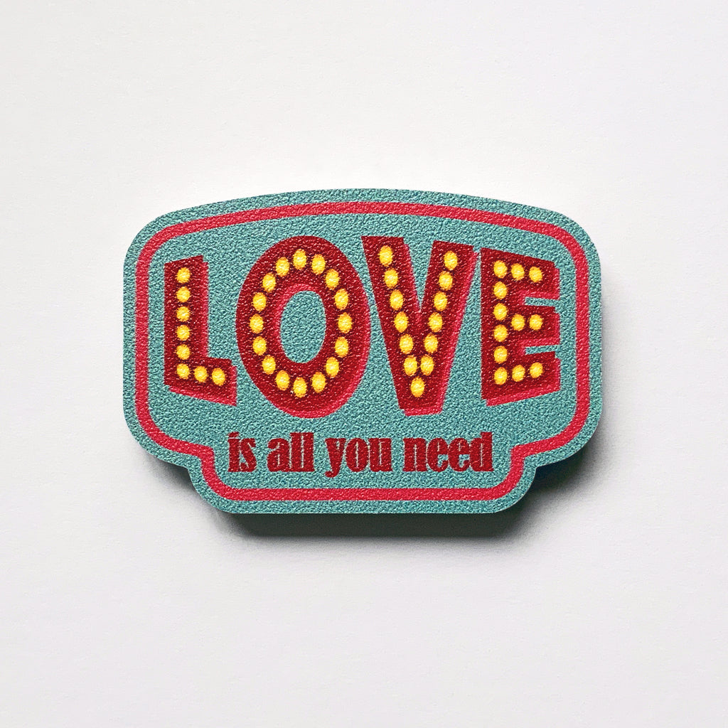 A vintage label shaped plywood fridge magnet by Beyond the Fridge with type that says love is all you need