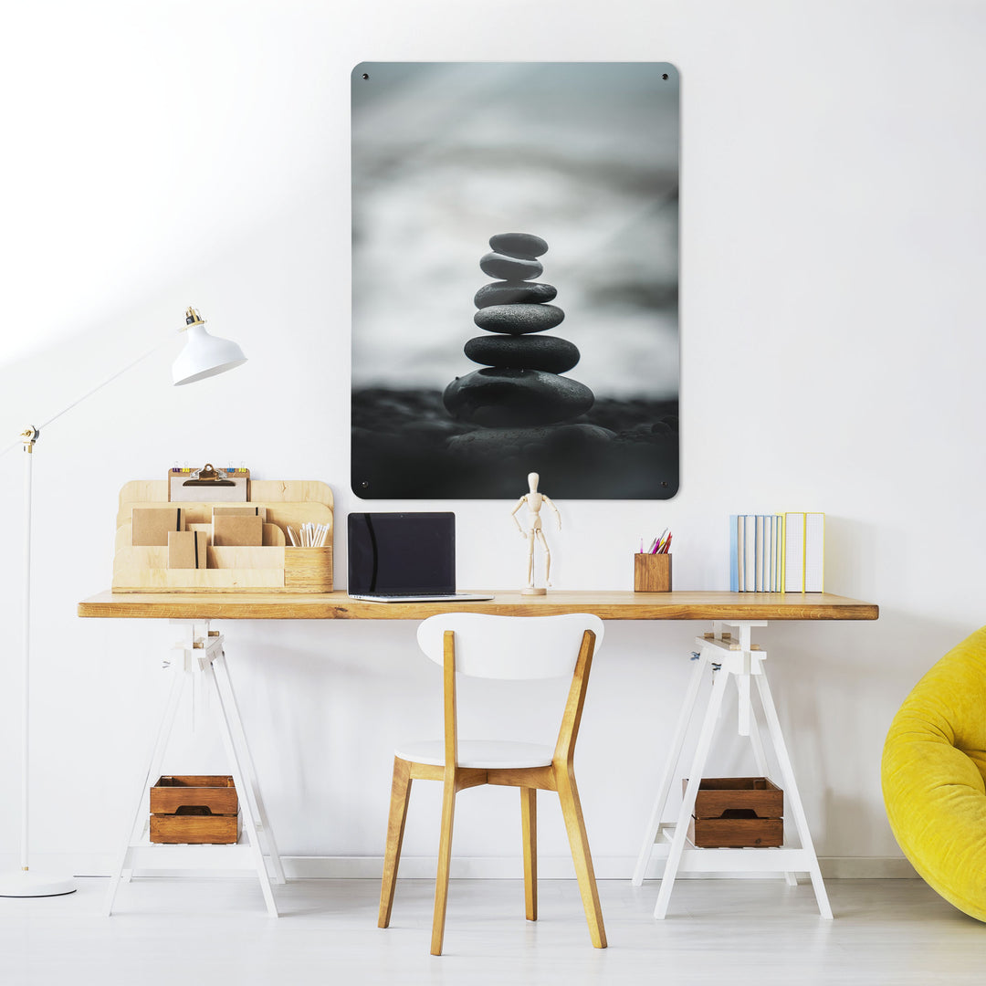 A desk in a workspace setting in a white interior with a magnetic metal wall art panel showing a photograph of a stack of balancing stones on a beach