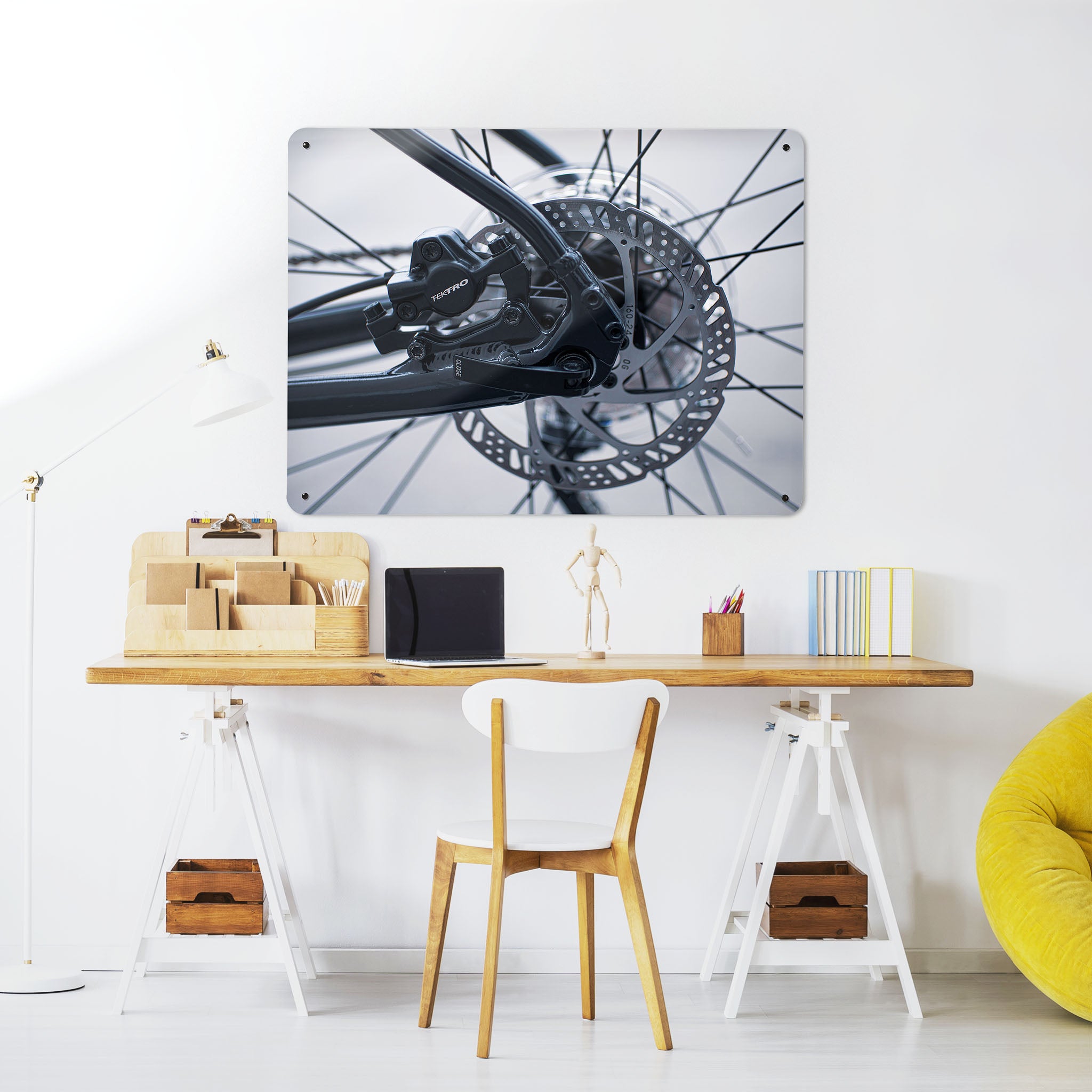 A desk in a workspace setting in a white interior with a magnetic metal wall art panel showing a black and white photograph of bicycle spokes