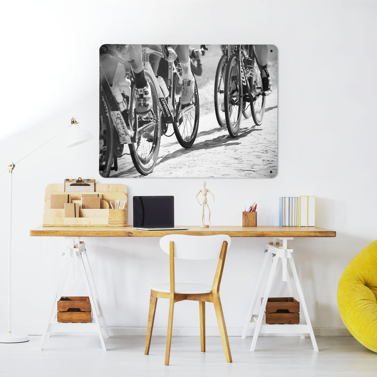 A desk in a workspace setting in a white interior with a magnetic metal wall art panel showing a black and white photograph of cyclists