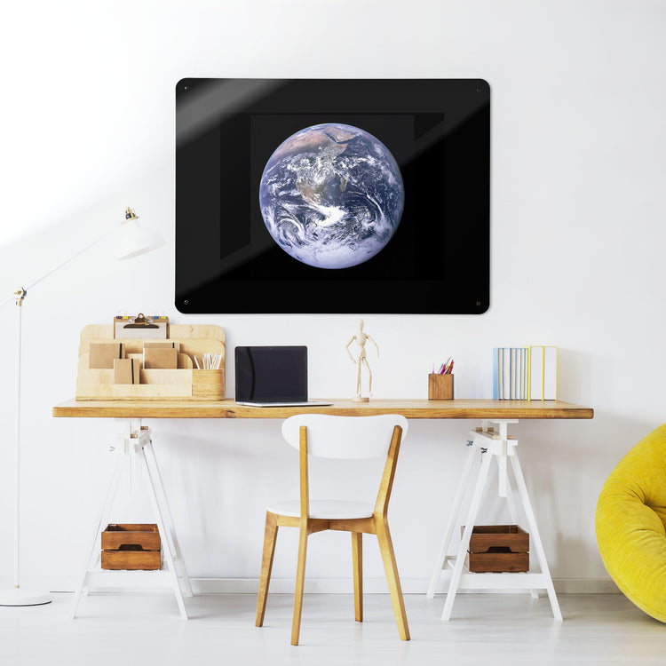 A desk in a workspace setting in a white interior with a magnetic metal wall art panel showing a photograph of planet Earth shot from space