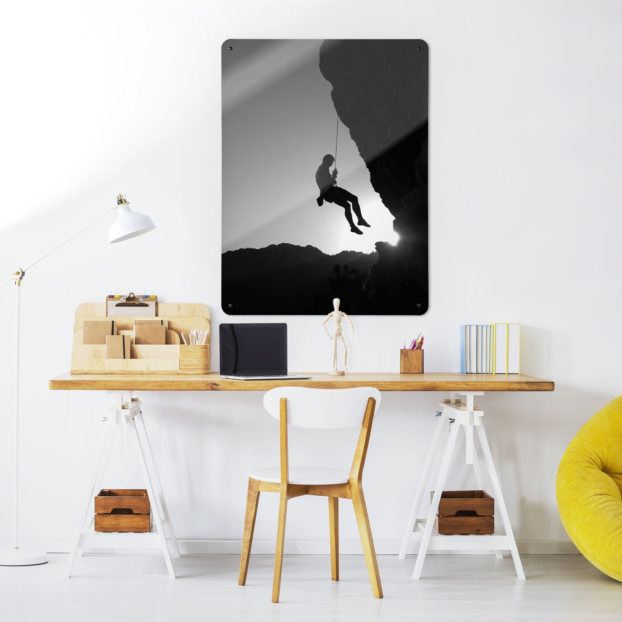 A desk in a workspace setting in a white interior with a magnetic metal wall art panel showing a black and white photographic image of a rock climber suspended on a cliff in silhouette 