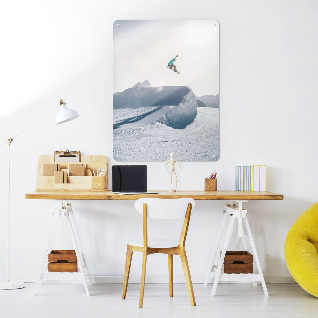 A desk in a workspace setting in a white interior with a magnetic metal wall art panel showing a photograph of a snowboarder jumping on a snowboard in a snowy landscape