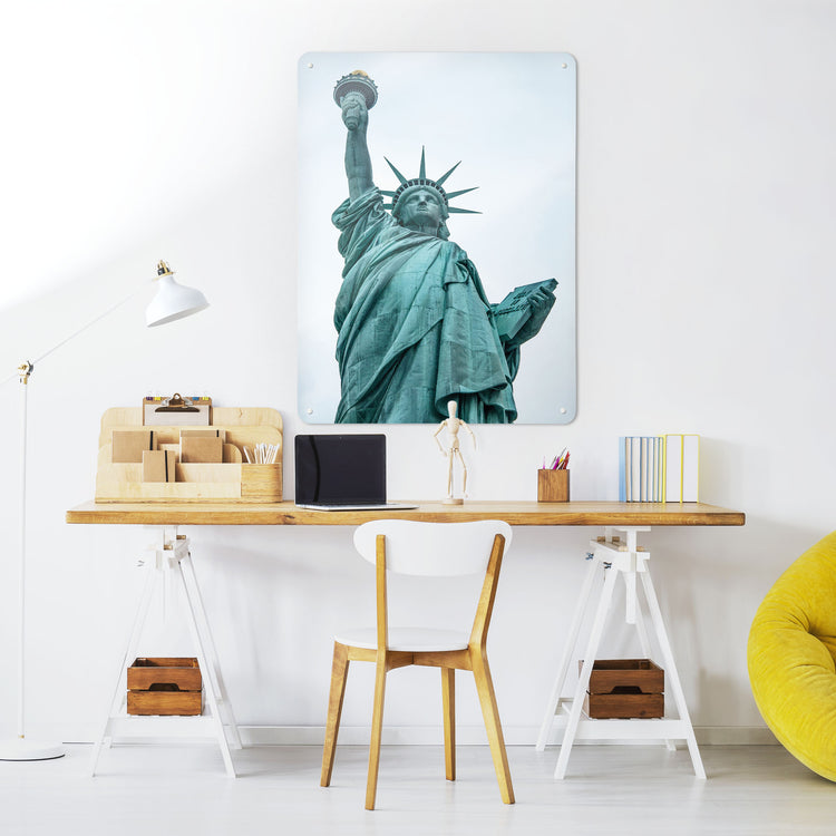 A desk in a workspace setting in a white interior with a magnetic metal wall art panel showing a photograph of the Statue of Liberty