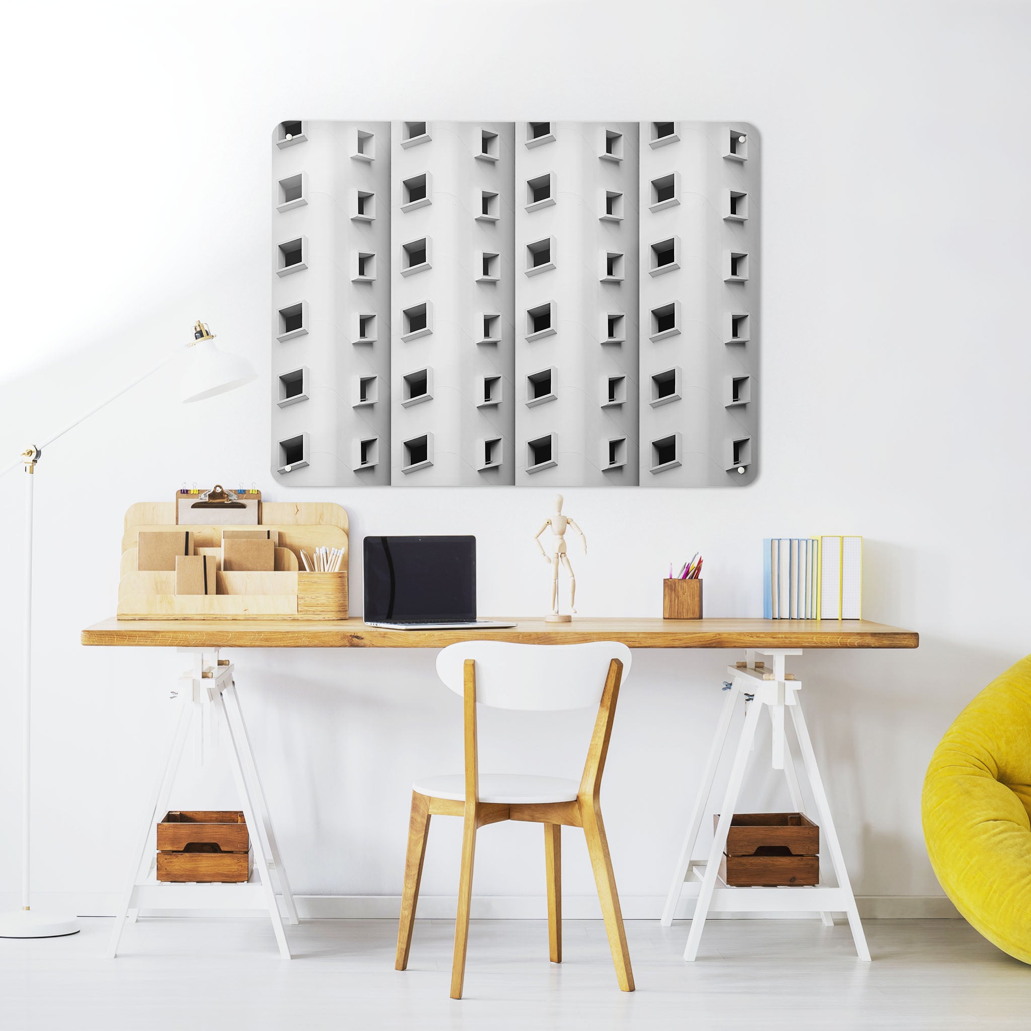 A desk in a workspace setting in a white interior with a magnetic metal wall art panel showing a black and white architectural photograph of a building with a pattern of windows