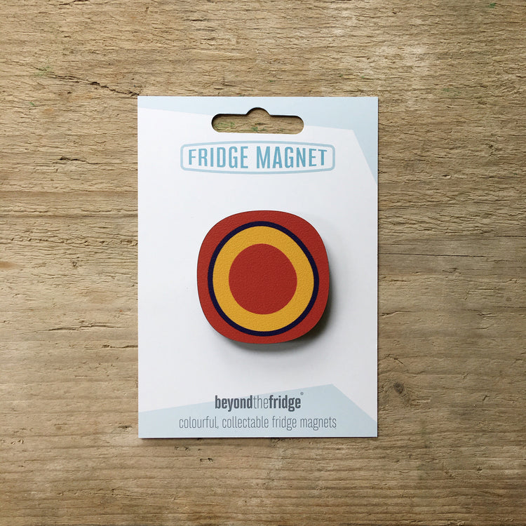 A red circle millefiori design plywood fridge magnet by Beyond the Fridge in it’s pack on a wooden background
