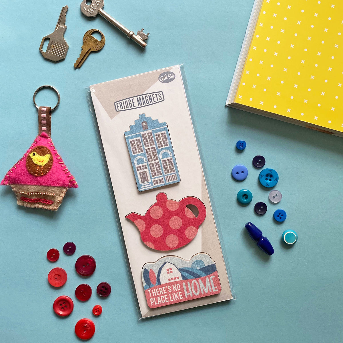 A blue table top with gift set of three Fridge Magnets for new home owners with Delft house, vintage label with there’s no place like home and pink teapot magnets - by Beyond the Fridge