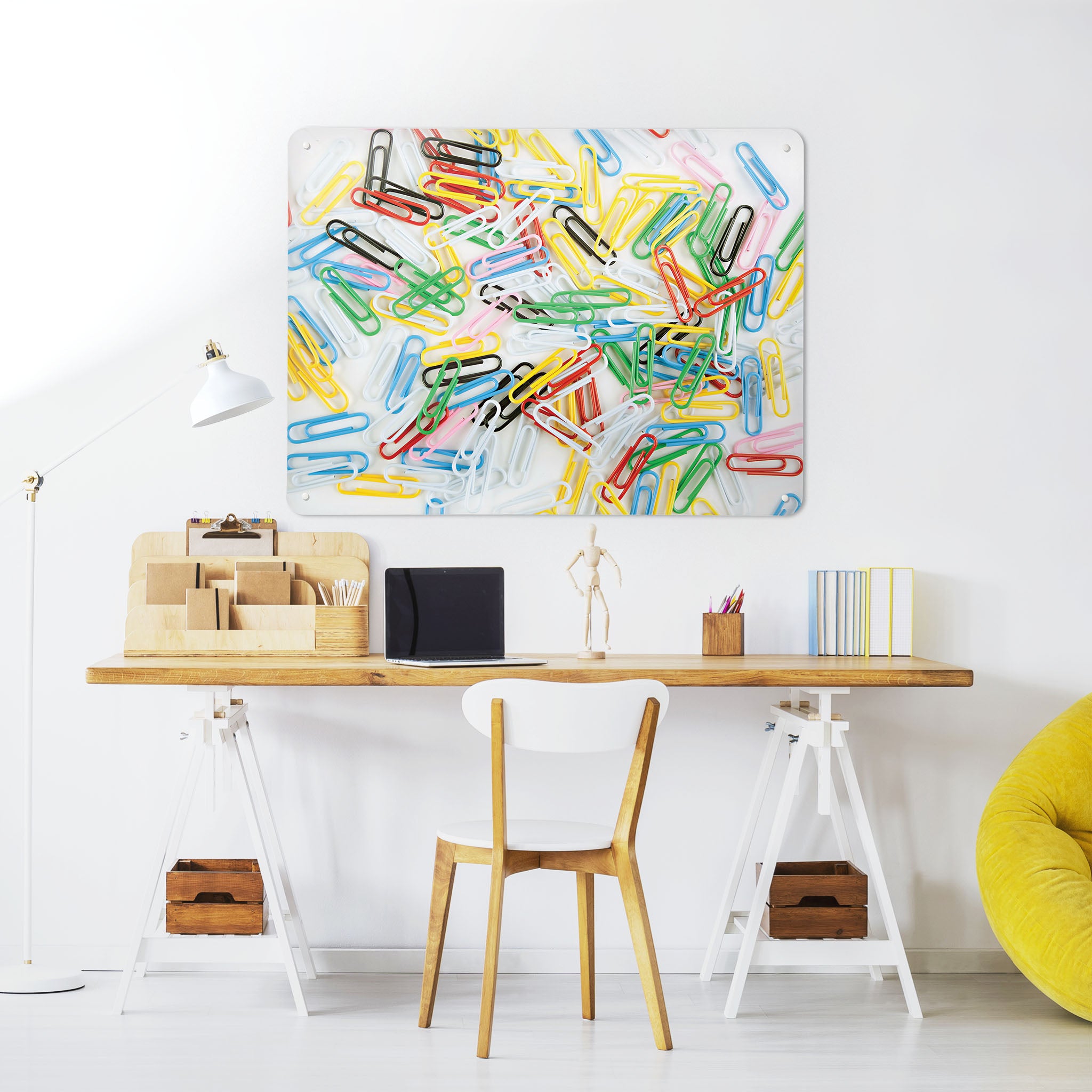 A desk in a workspace setting in a white interior with a magnetic metal wall art panel showing a photograph of multi coloured paper clips on a white background