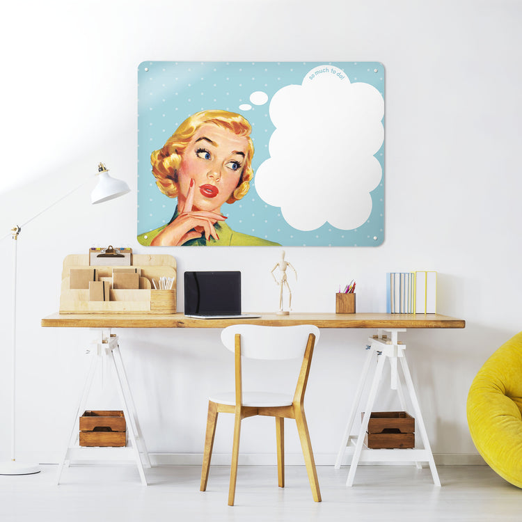 A desk in a workspace setting in a white interior with a dry wipe magnetic metal wall art panel showing a vintage girl illustration on a blue polkadot background and white thought bubble to write in