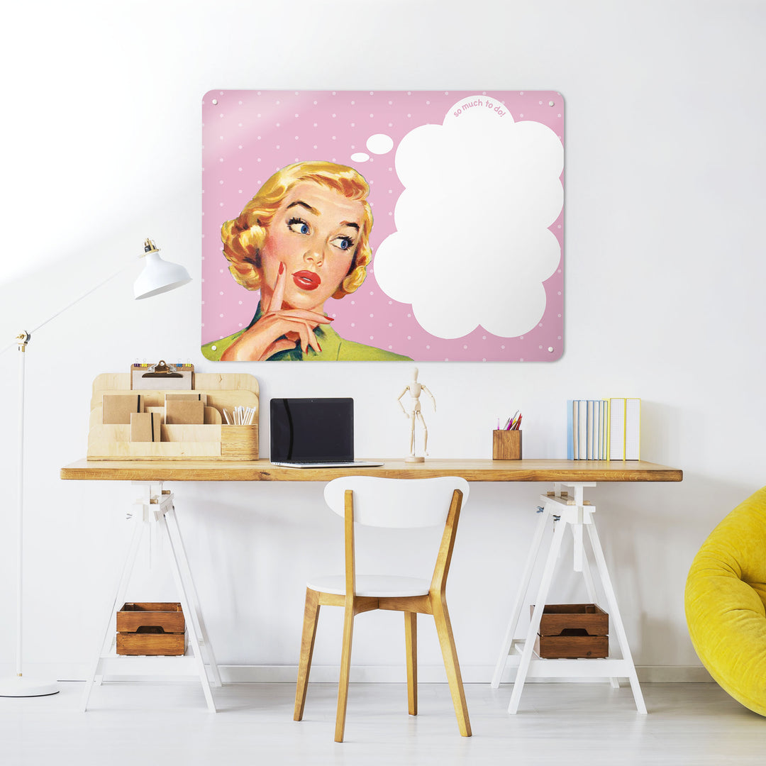 A desk in a workspace setting in a white interior with a dry wipe magnetic metal wall art panel showing a vintage girl illustration on a pink polkadot background and white thought bubble to write in