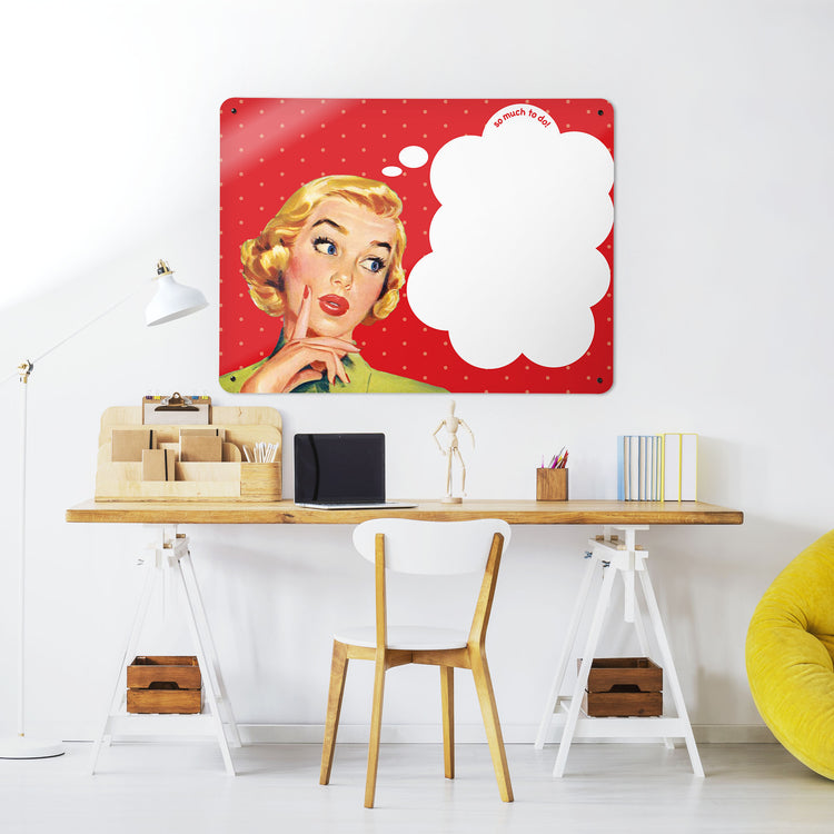 A desk in a workspace setting in a white interior with a dry wipe magnetic metal wall art panel showing a vintage girl illustration on a red polkadot background and white thought bubble to write in