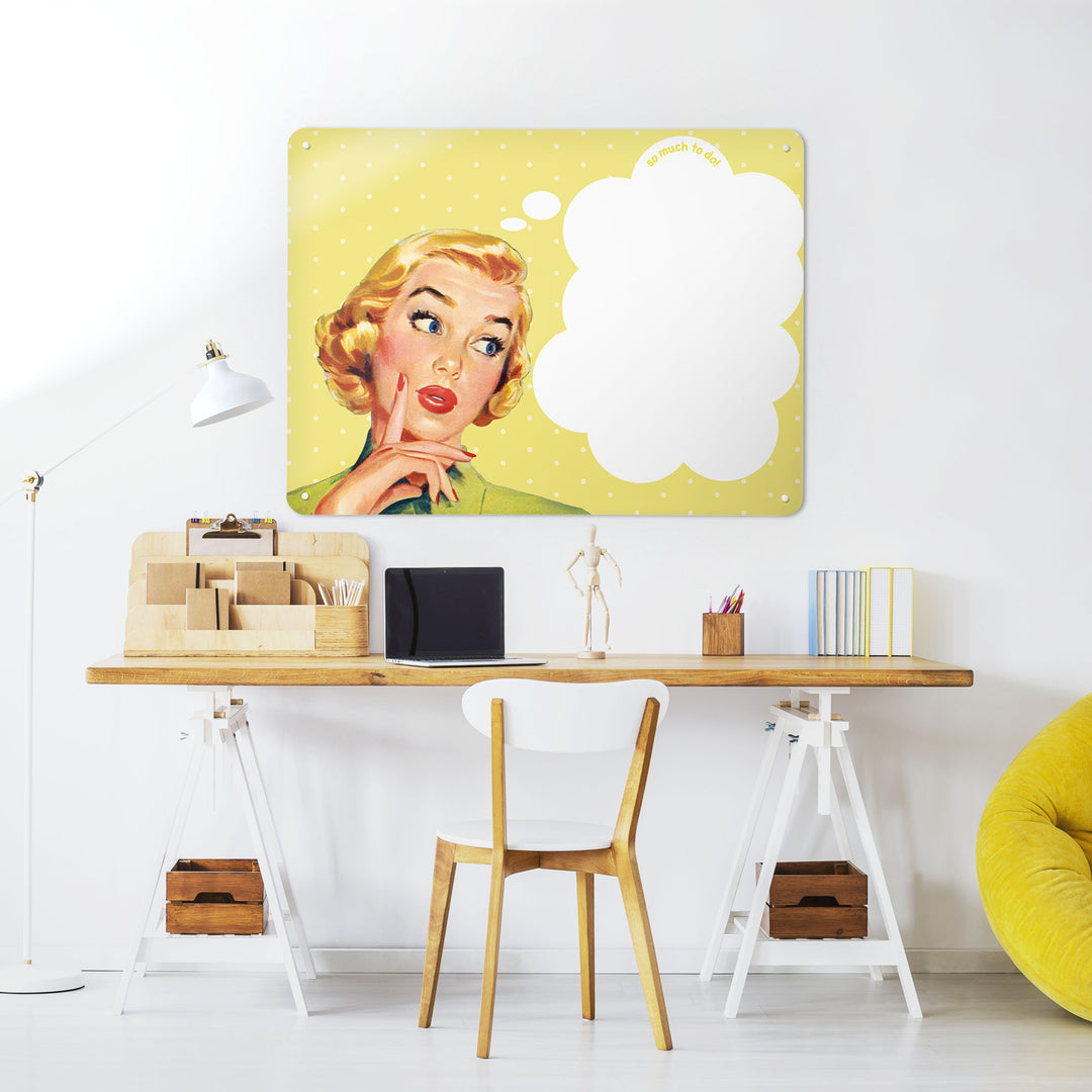 A desk in a workspace setting in a white interior with a dry wipe magnetic metal wall art panel showing a vintage girl illustration on a yellow polkadot background and white thought bubble to write in