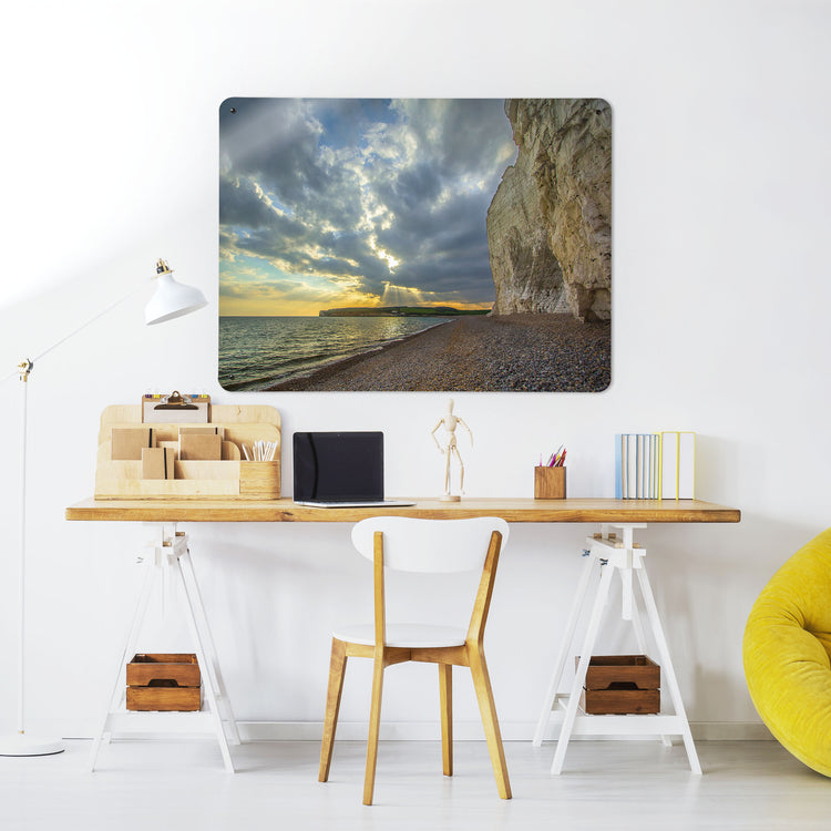 A desk in a workspace setting in a white interior with a magnetic metal wall art panel showing a photograph of cliffs and beach along the Sussex Coast at sunset