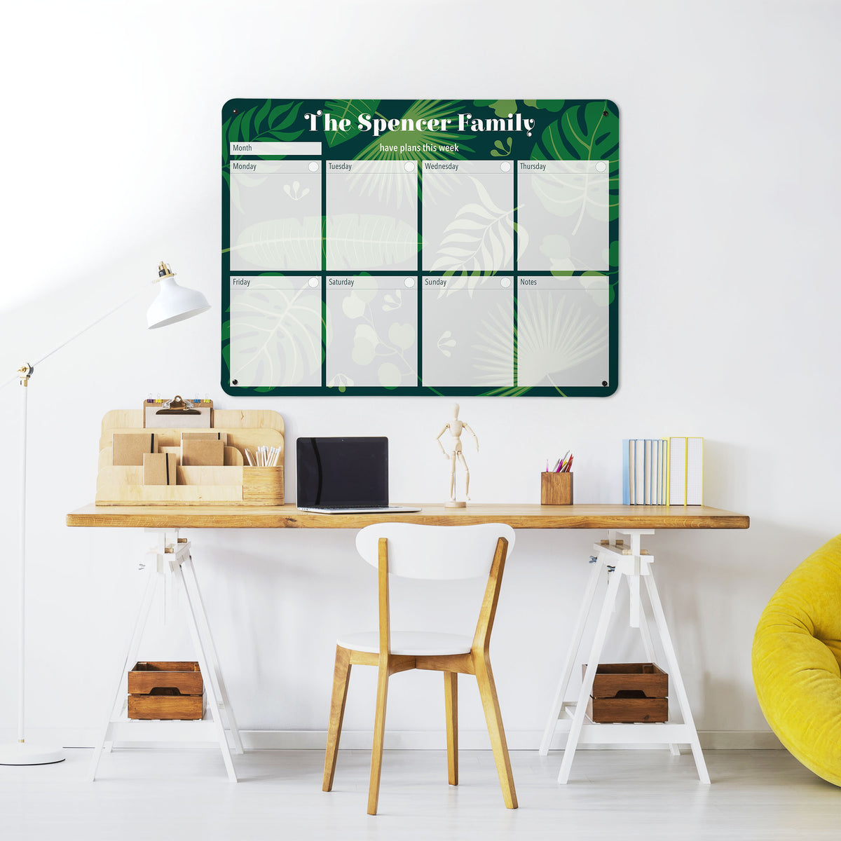 A desk in a workspace setting in a white interior with a magnetic metal wall art panel showing a tropical leaves design weekly planner to personalise