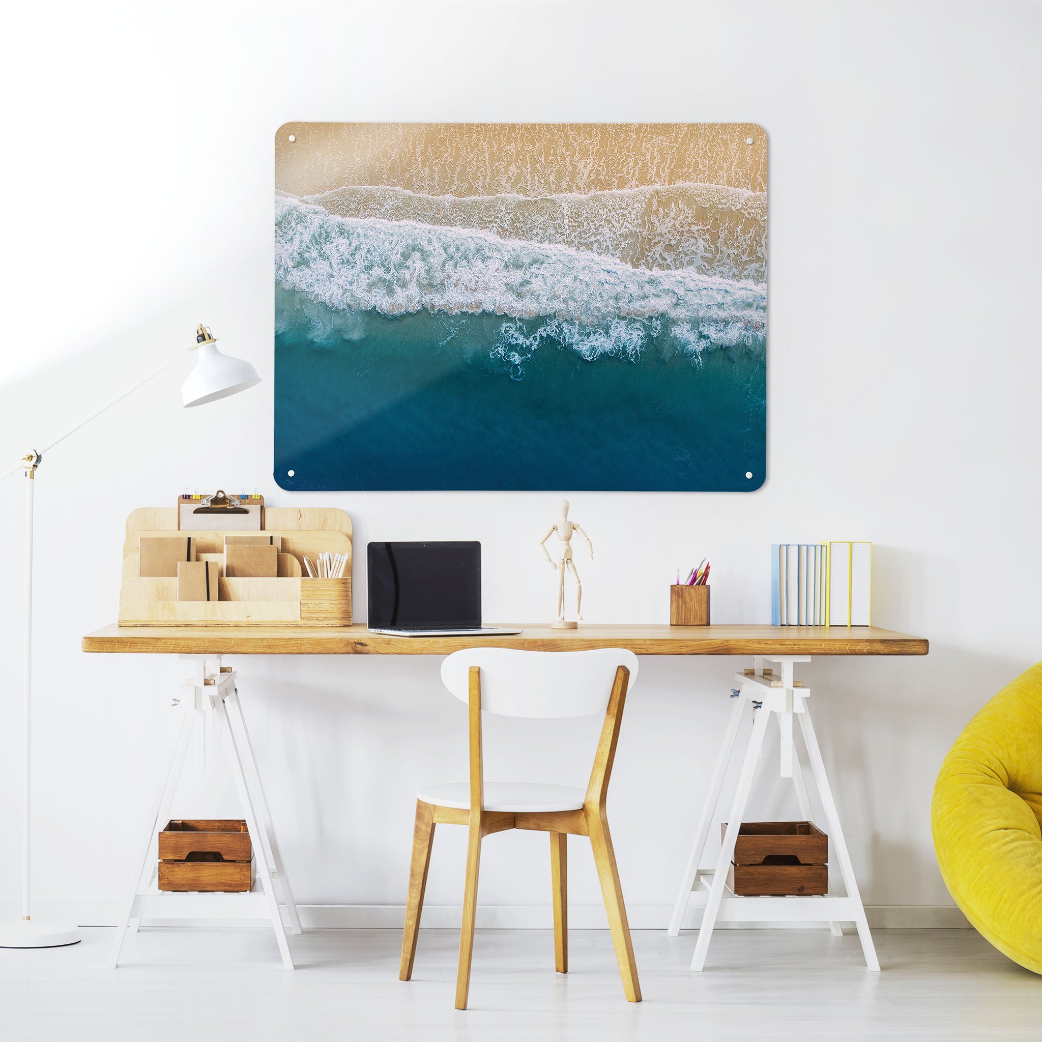 A desk in a workspace setting in a white interior with a magnetic metal wall art panel showing a photograph of wave breaking on a sandy beach