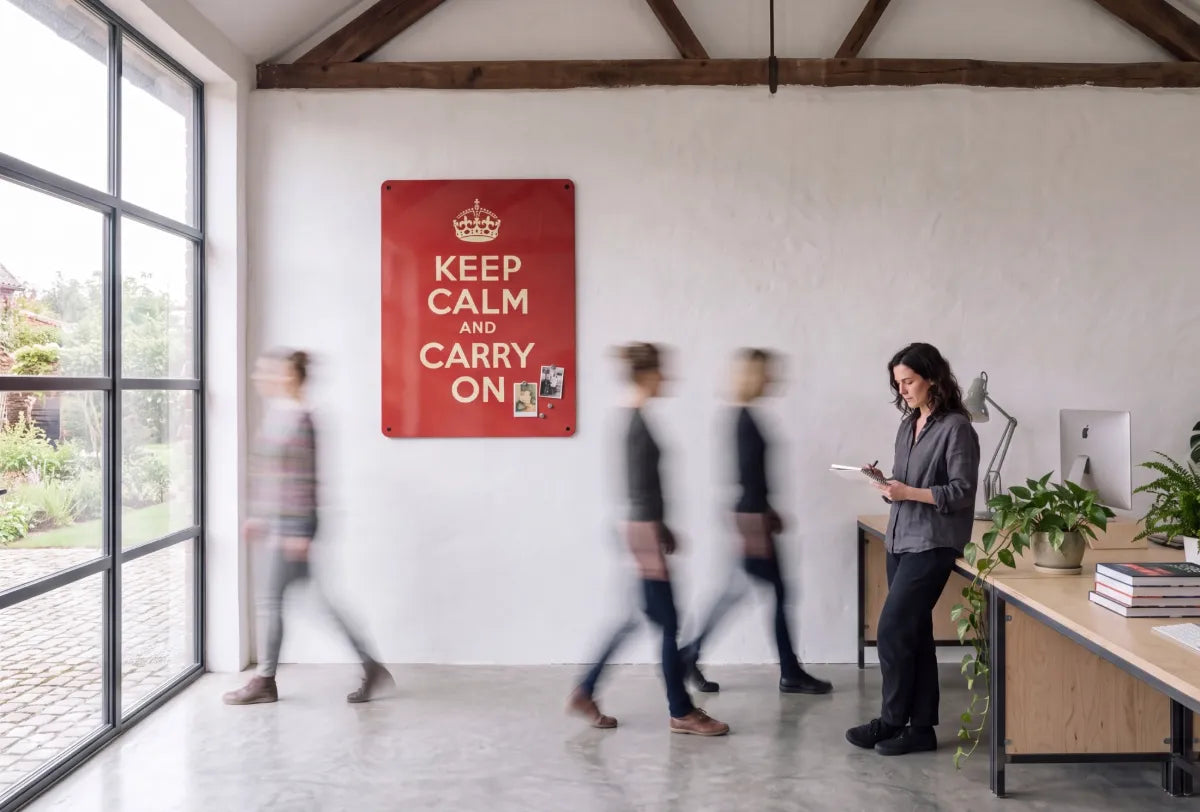 A large red Keep Calm and Carry On steel notice board on the whitewashed wall of a converted barn studio, with photographs held on it by magnets, a woman writing at a plywood desk and three figures blurred as they walk past