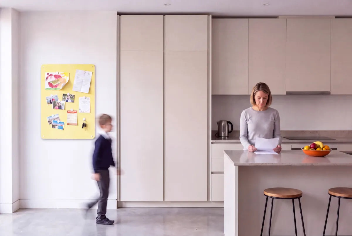 A plain pale yellow magnetic steel panel on the wall of a handleless integrated kitchen, holding a child’s painting, a school letter, photographs and keys, with a boy walking past and a woman reading at the island