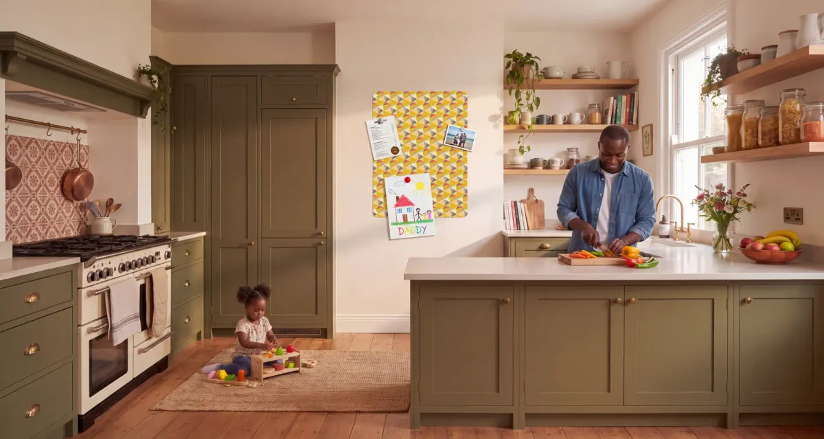 A family kitchen with olive Shaker units. A man chops peppers at the counter while his daughter plays on the floor. On the wall between them, a yellow geometric magnetic board holds a menu, a beach photograph and a child’s drawing signed Daddy.